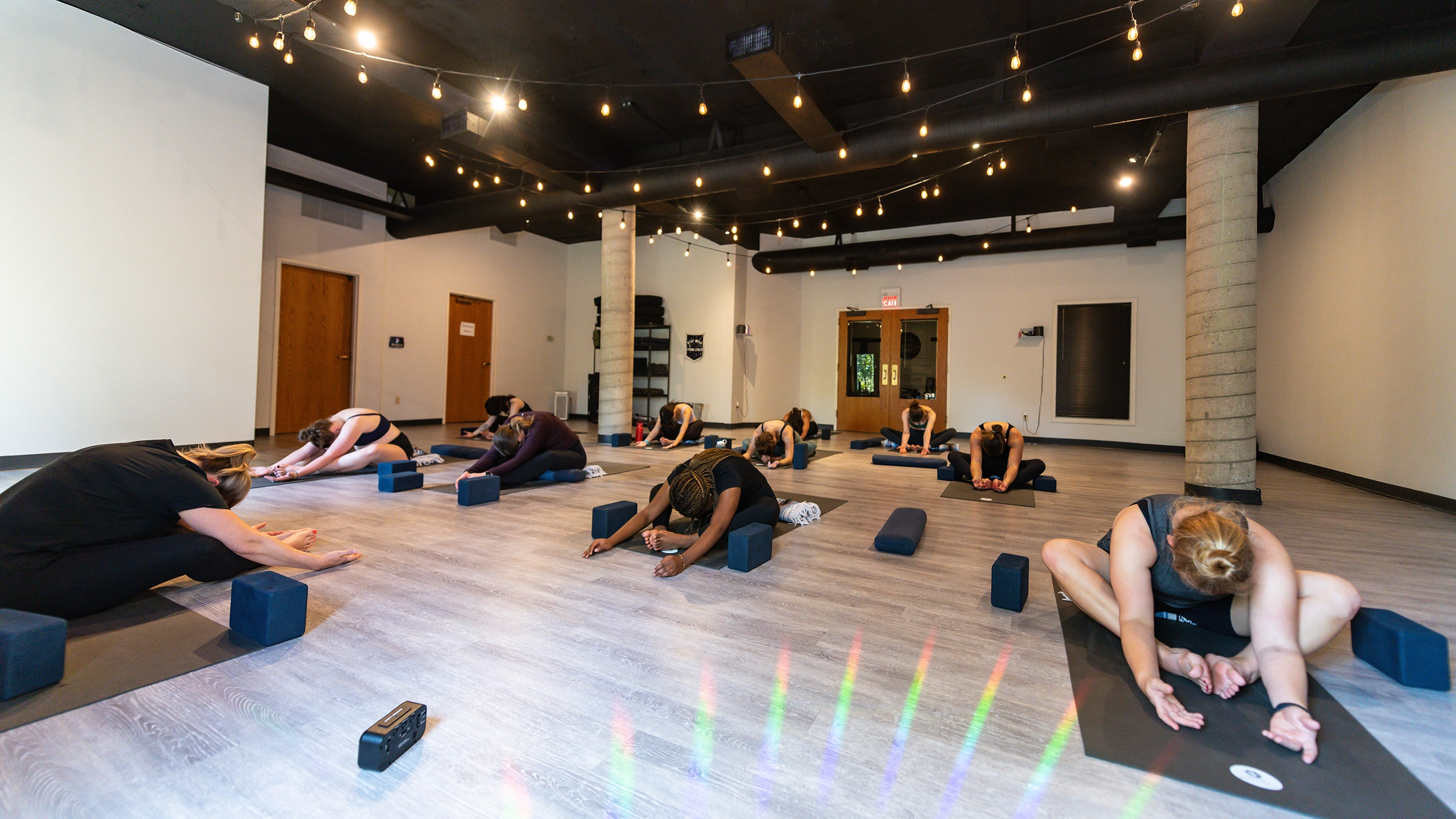 A diverse group of yoga students sit on mats folded over and spread out in a large studio with twinkle lights hanging along the ceiling.