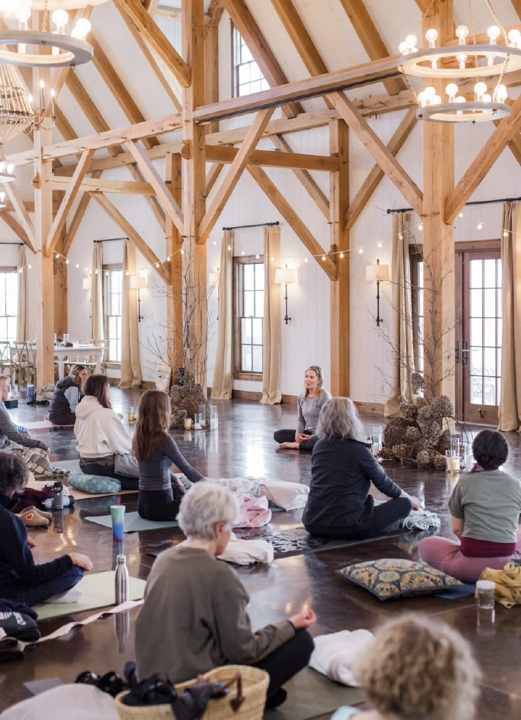 A large group of students are sitting on yoga mats in a large naturally lit room with oak timber architecture, white walls, dark wood floors, and decorated with natural elements. Pillows an dress of guests makes the room feel lived in and comfortable.