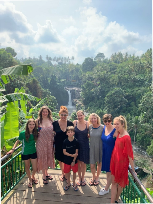 A small group of women an children gather for a posed photo infront of a lush forest and waterfall. 