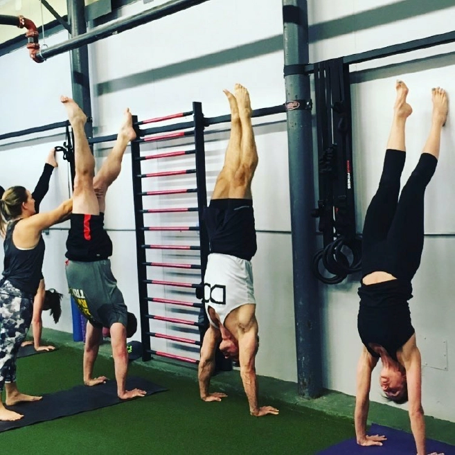 Yoga workshop students do handstands against a wall while receiving instruction in a strength gym.