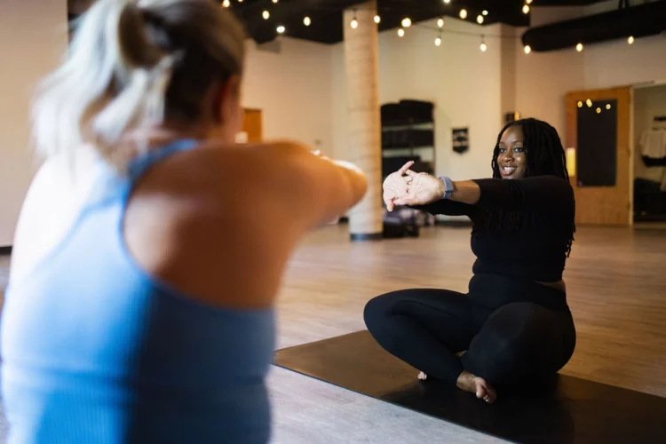 A private yoga instruction student stretches while sitting on a yoga mat in a studio. Sarah's back is to the camera, with the focus on a smiling student actively watching instructor Sarah Sandkuhl.